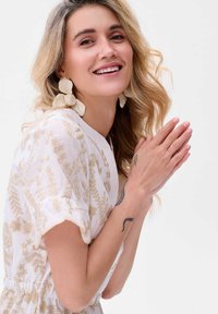 Witte jurk met gouden botanische print, korte mouwen, elastische taille; grote crèmekleurige oorbellen; licht, golvend blond haar; handen samengevoegd.