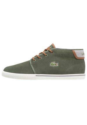 Zapatilla baja verde de ante con cordones marrones, suela de goma blanca y logo de cocodrilo verde bordado en el lateral.
