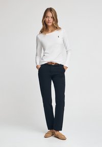 Biała koszula z długim rękawem z małym logo, połączona z ciemnoniebieskimi spodniami i brązowym paskiem. Model nosi beżowe loafersy. Prosty, dopasowany krój.