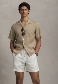 Beige short-sleeve shirt in linen fabric with collar and pocket; paired with white drawstring shorts; black sunglasses and blue watch detail.