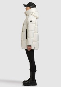 Biela páperová bunda s vysokým golierom a čiernym bočným zipsom. Kombinovaná s čiernymi nohavicami a mohutnými čiernymi topánkami.