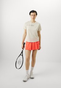 Sporty antrekk med en lys beige kortermet skjorte med en subtil logo og lys orange shortser, kombinert med hvite joggesko og et tennisrack.