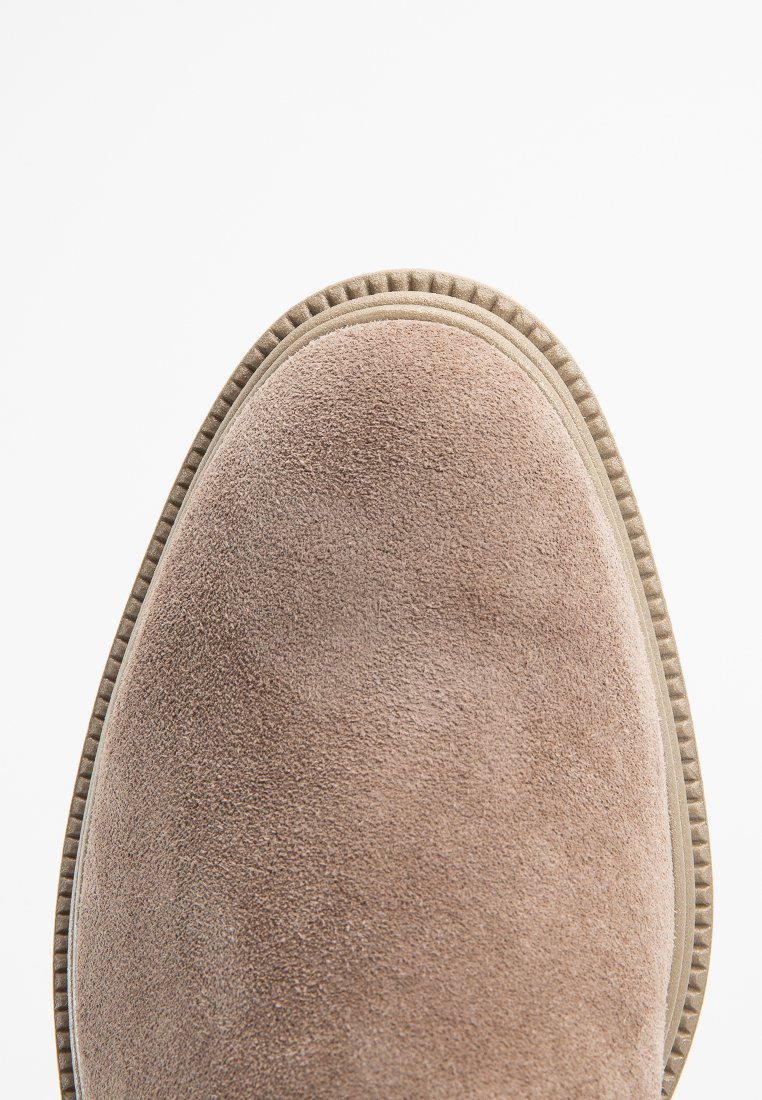 Beżowe zamszowe buty z zaokrąglonym noskiem, teksturowaną powierzchnią i kontrastowym szwem wzdłuż podeszwy.