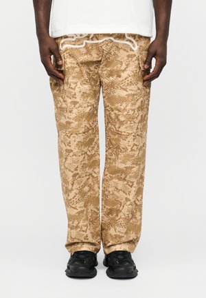 Persoon die tankleurige camouflage cargobroek, zwarte sneakers en een wit shirt draagt, staand tegen een effen witte achtergrond.