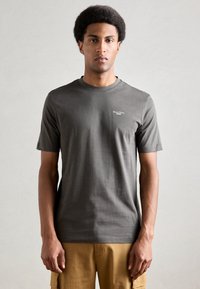 Pelēks kokvilnas T-krekls ar īsām piedurknēm, apaļo kakla izgriezumu un diskrētu logo, kas iespiests kreisajā krūtī, uz neitrāla fona.