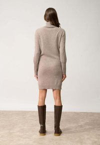 Abito maglione in beige chiaro, con scollatura alta e maniche lunghe, abbinato a stivali al ginocchio lucidi marroni.