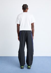 Black lightweight pants with navy side panels, elastic waistband, and a relaxed fit. Paired with a plain white t-shirt and green shoes.