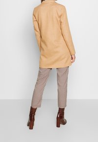 Persona vestida con un abrigo largo color camel, pantalones de cuadros hasta el tobillo y botines de tacón marrón, de pie contra un fondo liso.