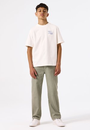T-shirt en coton beige clair à manches courtes avec un imprimé graphique sur la poitrine, associé à un pantalon en velours côtelé vert clair et des baskets blanches.