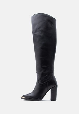 Black leather knee-high boot with pointed toe, gold metal tip, and block high heel on white background.