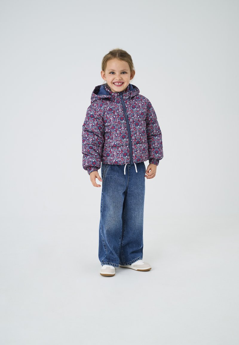 Ung pige smiler, iført en mønstret dunjakke med hætte, brede blå jeans og hvide sneakers, stående på en ensartet lys baggrund.