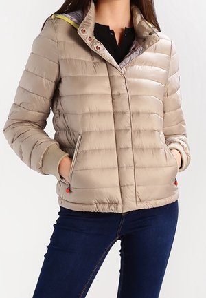 Vrouw draagt taupekleurige doorgestikte pufferjas met zijritssluitingszakken over een zwarte top en donkerblauwe jeans, handen in de zakken.