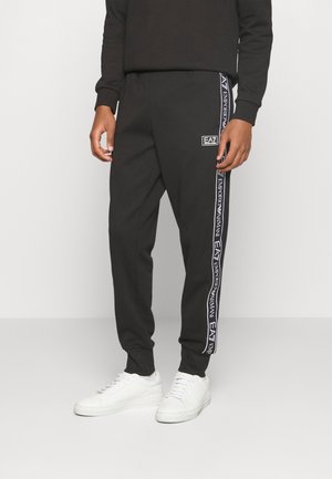 Pantalon de survêtement noir avec des bandes blanches au logo EA7 Emporio Armani sur les côtés, porté avec des baskets blanches par une personne debout.