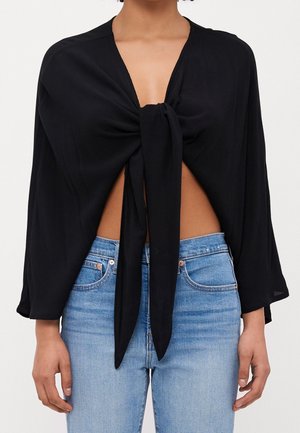 Femme portant un haut noir à manches longues noué devant, au-dessus du ventre, associé à un jean bleu clair taille haute.