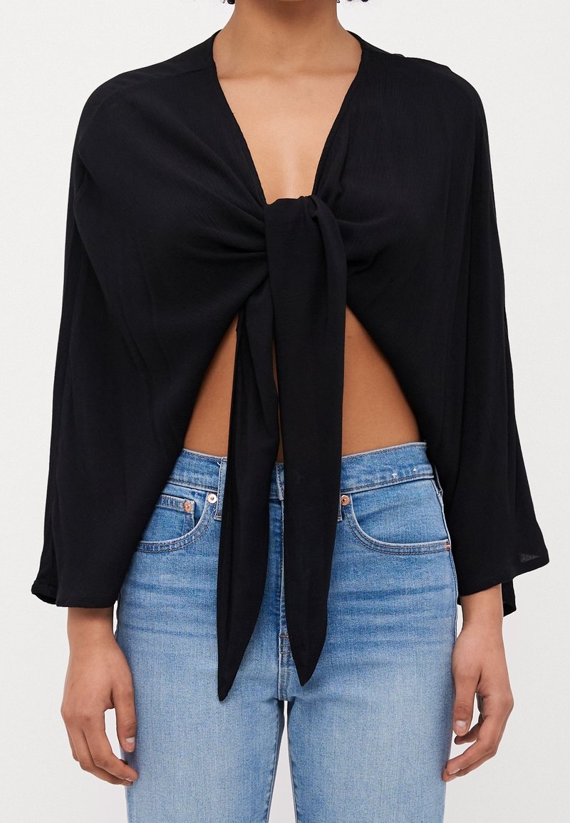 Femme portant un haut noir à manches longues noué devant, au-dessus du ventre, associé à un jean bleu clair taille haute.