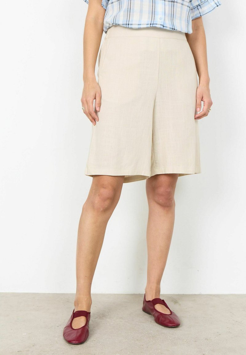 Personne portant un short beige jusqu'aux genoux, une chemise à carreaux bleue rentrée, et des chaussures plates rouges, debout sur un sol clair contre un mur blanc.