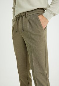 Beige Jogginghose mit Zugbandbund, seitlichen Taschen und einer strukturierten Stoffoberfläche. Sie hat einen dezenten Glanz und kontrastierende Seitennähte.