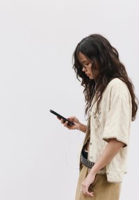 Cămașă bej, texturată, cu un model decorativ; pantaloni verzi măslinii; ținând un smartphone cu căști; păr lung și ondulat.