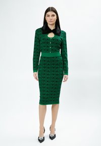 Grünes Kleid mit schwarzem geometrischem Muster, langen Ärmeln, Schlüsselloch-Ausschnitt und Kragen aus Kunstfell. Enganliegende Form, knielang, kombiniert mit schwarzen Absätzen.