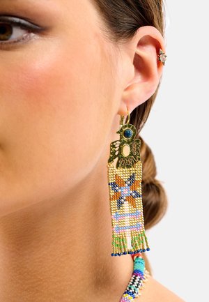Les boucles d'oreilles dorées présentent un motif d'oiseau en haut, avec une frange de perles colorées en bleu, orange et vert, mettant en valeur des motifs et des textures complexes.