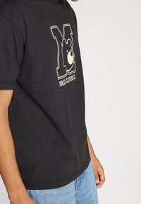 Černé bavlněné tričko s odvážnou bílou grafikou písmena "M" a obličejem medvěda, s textem "M.A.I.A Clothing Co." pod ním. Krátké rukávy.