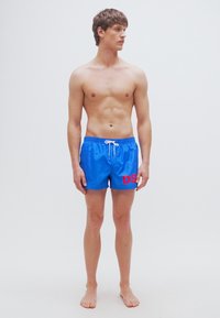 Μπλε μαγιό σορτς με κορδόνι στη μέση, με κόκκινο λογότυπο "DS" στην αριστερή πλευρά, κατασκευασμένο από λείο, ελαφρύ ύφασμα.