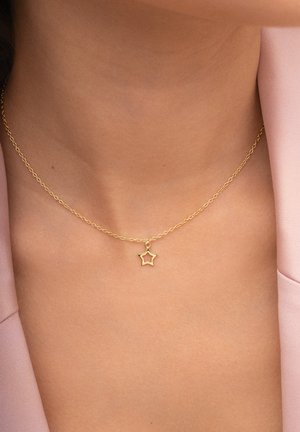 Collier en chaîne en or avec un petit pendentif en forme d'étoile, porté sur un cou nu devant un fond en tissu rose. Design simple et texture lisse.
