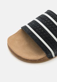 Svart sandale med to teksturerte stropper med hvite striper. Sålen er naturlig tan, noe som gir en kontrast til det sorte overdelen.