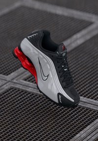 Melna un pelēka Nike sporta kurpe ar sarkaniem pavasara tipa zoles elementiem, novietota uz metāla režģa virsmas.