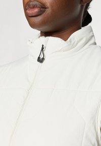 Bijela jakna od quilted materijala s visokim ovratnikom, zatvaranjem na zip, i detaljem crnog povlačnog užeta. Tkanina ima glatku, laganu teksturu.