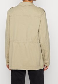 Jasno beżowa kurtka z kołnierzem, guzikami ozdobnymi i taliowaniem. Wykonana z wytrzymałej tkaniny, posiada dwa pagony na ramionach.