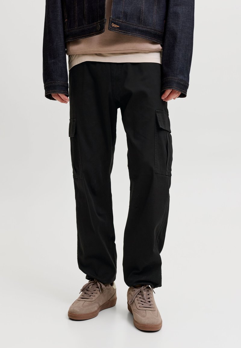 Personne portant un pantalon cargo noir, des baskets en daim beige, une chemise beige et une veste en jean sombre, debout sur un fond uni.