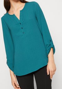 Blouse à manches longues couleur sarcelle avec col en V et patte de boutonnage, présentant des manches retroussées avec des accents de boutons et un tissu doux et texturé.