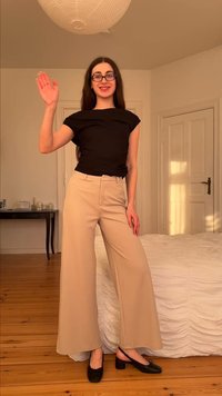 Γυναίκα με μακριά σκουρόχρωμα μαλλιά και γυαλιά, που κουνάει το χέρι, φοράει μαύρο τοπ, μπεζ φαρδιά παντελόνια και μαύρα παπούτσια σε υπνοδωμάτιο με ξύλινο πάτωμα.