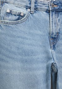 Gros plan sur un jean en denim bleu clair montrant les poches avant, les passants de ceinture, la fermeture par bouton et les détails de couture orange.