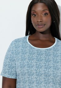 Mujer con cabello largo y oscuro, lleva pendientes de aro plateados y una camisa de manga corta azul con estampado blanco, expresión neutral, fondo blanco.