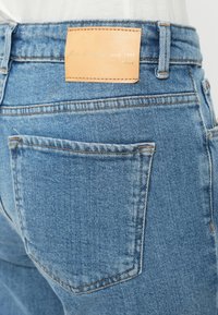 Μπλε τζιν με ελαφρώς φθαρμένη υφή, με δερμάτινο ταμπελάκι σε καφέ απόχρωση στη ζώνη της μέσης στο πίσω μέρος και πίσω τσέπη με διπλό ράψιμο.