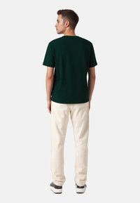 Dark green cotton t-shirt with short sleeves, paired with light cream relaxed-fit pants and black and white sneakers, viewed from behind.
