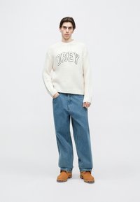 Pulover crem tricotat cu textul „OBEY” negru, asortat cu blugi albaștri largi și saboturi maro, stând pe un fundal alb.