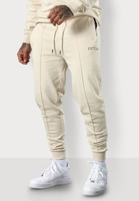 Cremefarbene Jogginghosen aus weichem Material, mit Seitentaschen, Kordelzugbund und gesteppten Details. Kombiniert mit weißen Turnschuhen.