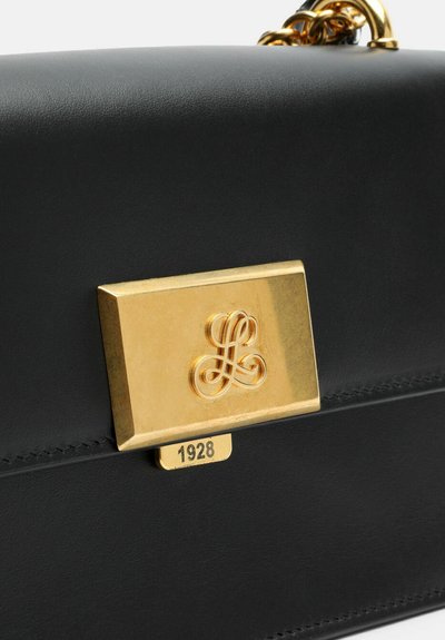 Melna ādas soma ar zelta toņa aizdari, uz kuras ir gravējums ar logotipu un detaļu "1928", gluda tekstūra un taisnstūrveida forma.