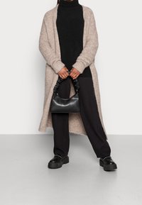 Cardigan beige en tricot porté sur un col roulé noir, assorti à un pantalon noir et des chaussures à plateformes. Un petit sac noir avec une poignée froncée est tenu en main.