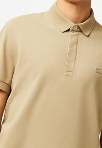 Beige polo tričko s texturovanou látkou, krátkými rukávy a dvoupanelovým zapínáním. Na hrudi má malé logo.