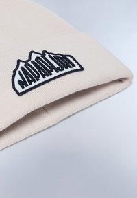 Knit beige beanie, jossa on musta brodeerattu pata, jossa on vuoristomotiivi ja teksti. Pehmeä rakenne ja taitos resorissa.