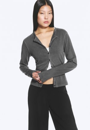 Junge Frau mit langen schwarzen Haaren trägt eine taillierte, dunkelgraue, geknöpfte Strickjacke über einem weißen Top und lockere schwarze Hosen, posiert mit verschränkten Händen.