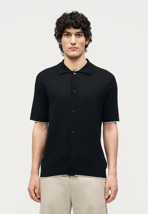 Junger Mann mit lockigem Haar, der ein schwarzes kurzärmeliges Hemd und hellbeige Hosen trägt, steht vor einem schlichten weißen Hintergrund.