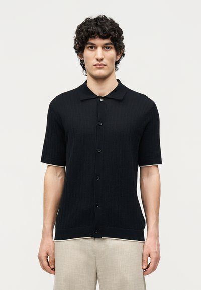 Jeune homme aux cheveux bouclés portant une chemise noire à manches courtes boutonnée et un pantalon beige clair, debout devant un fond blanc uni.