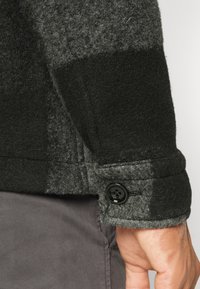 Primer plano de la muñeca de una persona que lleva una chaqueta de lana a cuadros negra y gris con un puño negro abotonado sobre pantalones grises.