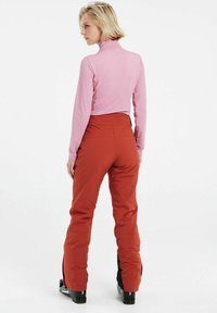 High-neck pink long-sleeve top paired with orange ski pants. The pants feature a high waist and ankle zippers; sleek black ski boots complete the outfit.