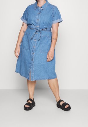 Persoon draagt een blauwe denim jurk met knopen, een gestrikte tailleband, opgerolde korte mouwen, grote voorzakken en zwarte stevige sandalen.